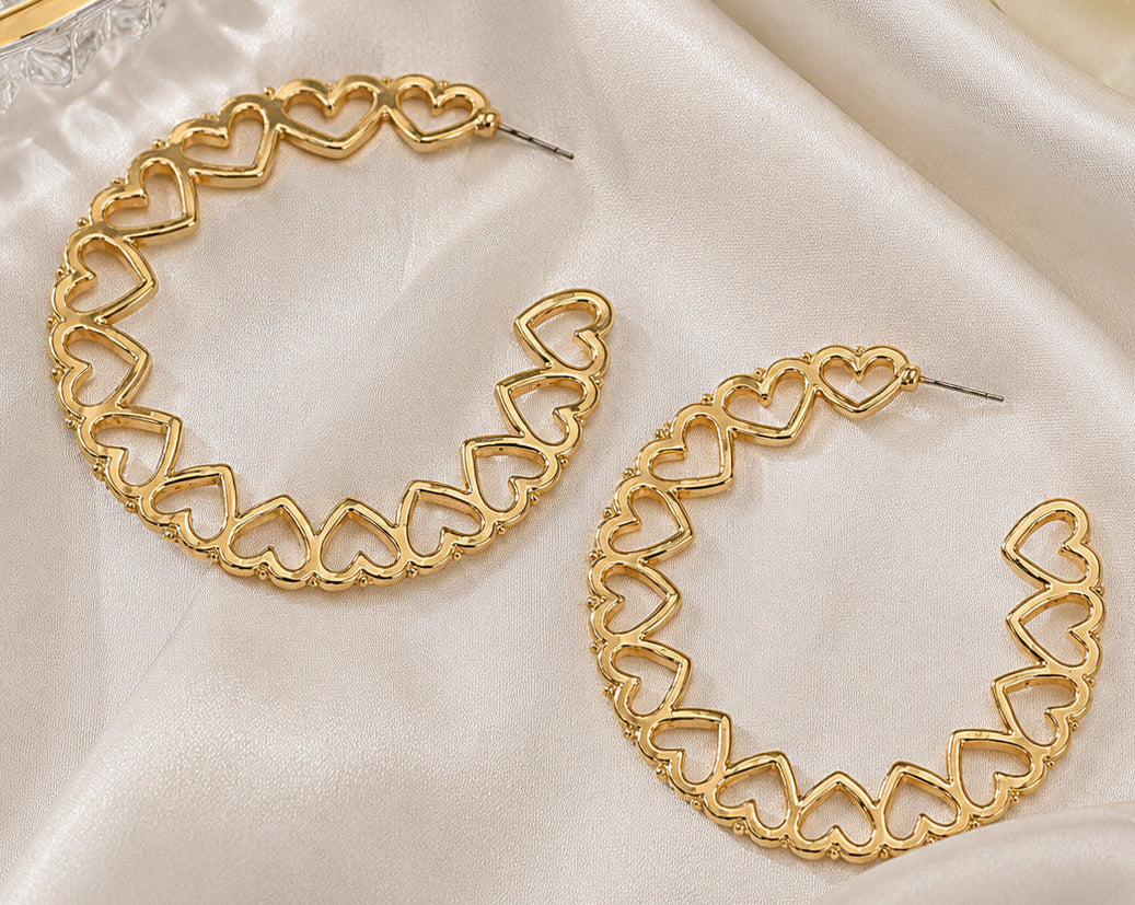 A pair of gold hoop earrings with an openwork heart-link design, displayed on draped white satin fabric next to a white rose and a glass dish.