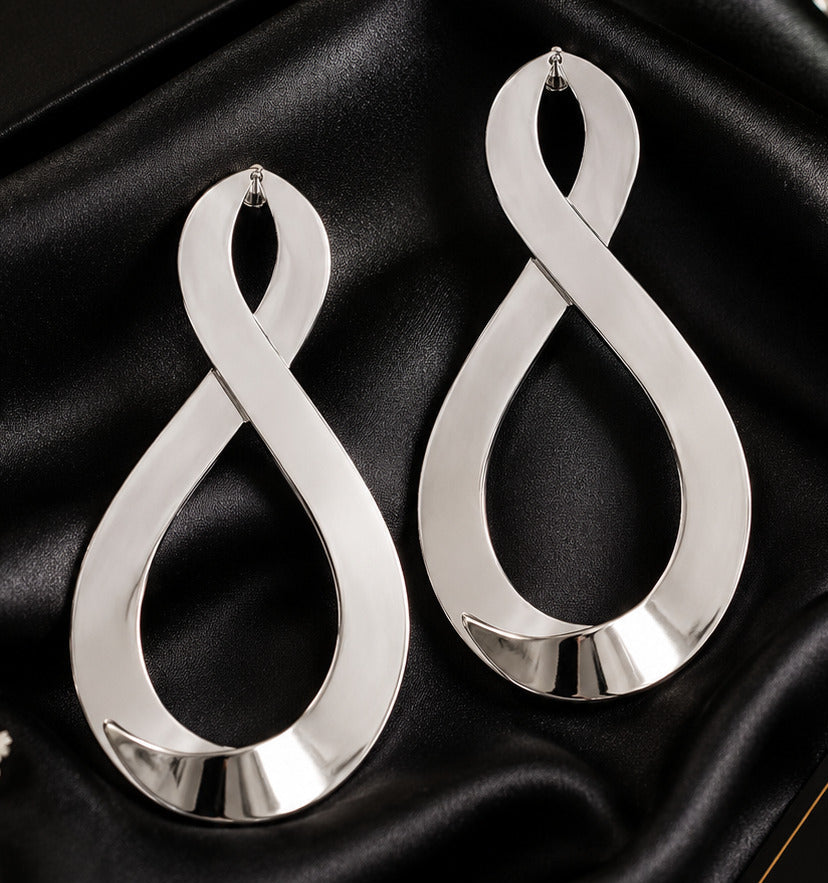 A pair of large silver infinity-shaped hoop earrings displayed on black satin fabric, with a black Rubylee Elegance box and small white flowers in the background.