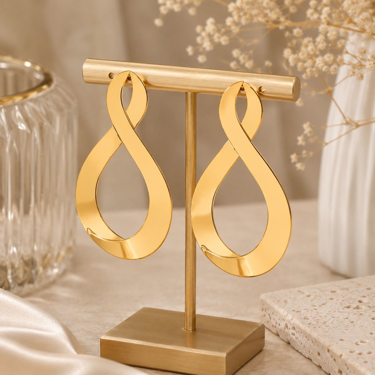A pair of large gold infinity-shaped hoop earrings displayed on a matching gold stand, set against a softly styled neutral background with dried flowers and glassware.