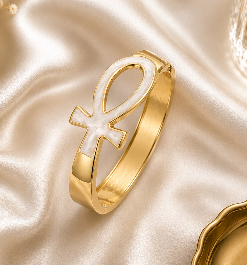 A gold-toned cuff bracelet featuring a large white inlay of the ankh symbol, styled on draped champagne satin with dried flowers and a glass bottle in the background.