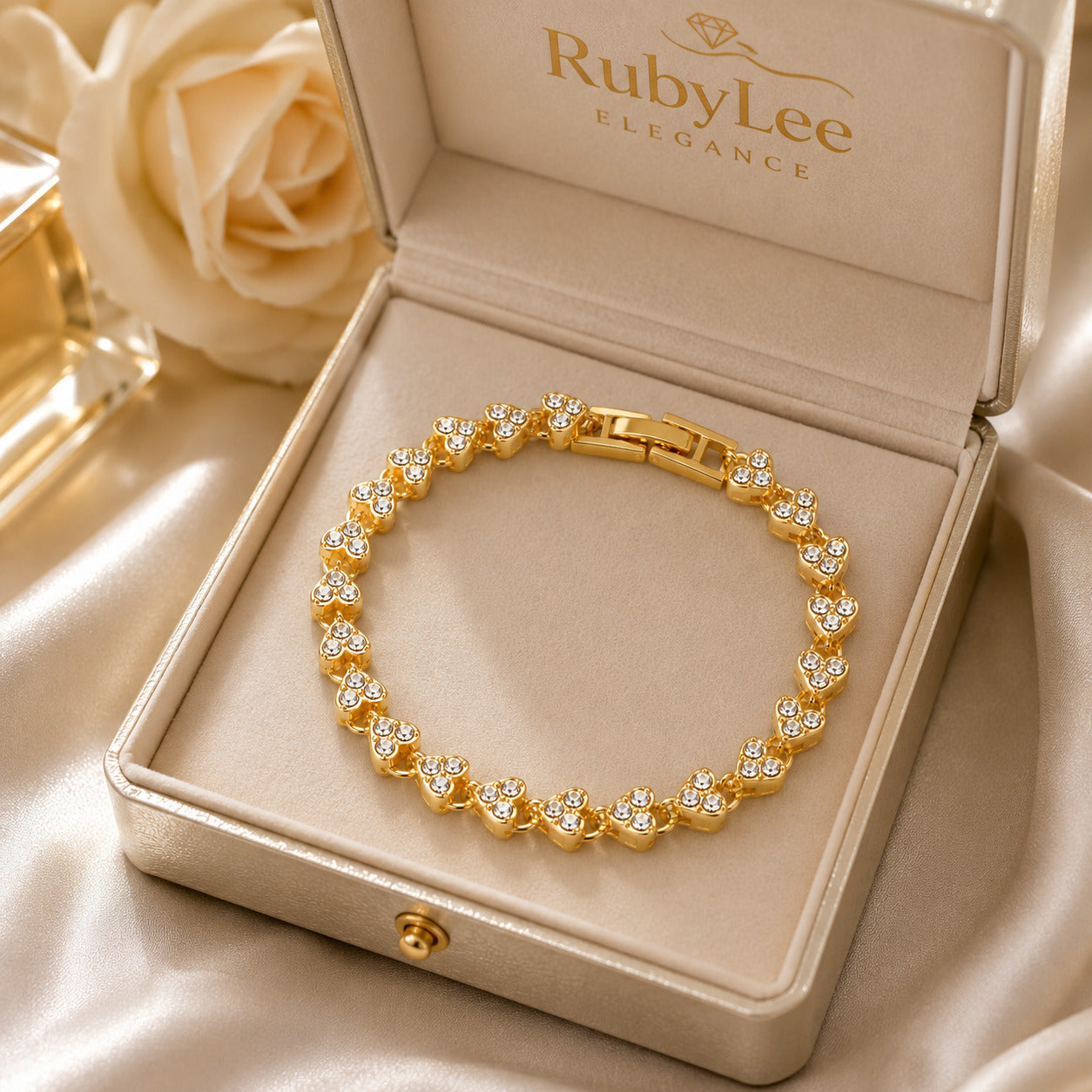 A gold bracelet with heart-shaped clear stones displayed inside an open RubyLee Elegance jewelry box on a champagne satin background with a rose prop.