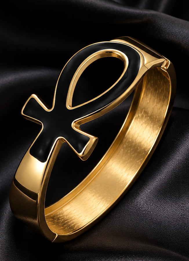 A gold-toned bracelet featuring a large black ankh symbol inlay, displayed on a draped black satin fabric background.
