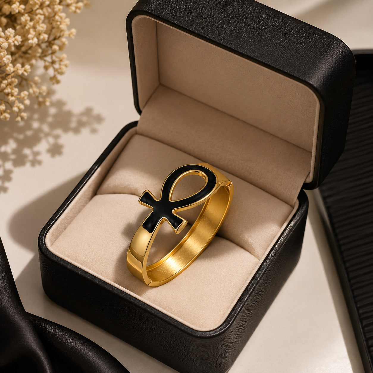 A gold bracelet with a black ankh symbol on the front, displayed inside an open black jewelry box with a beige velvet lining.