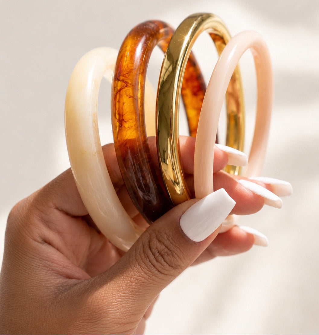 A hand with white manicured nails holds four smooth bangles in different colors: two translucent amber-brown, one polished gold, and one pale blush pink, against a softly lit light background.