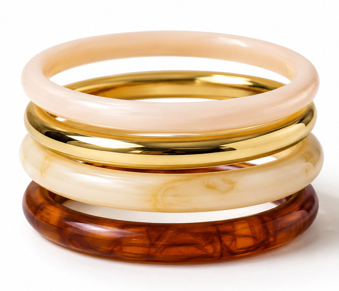 Four stacked bangles on a white background: a pale blush pink translucent bangle, a polished gold bangle, a cream marbled bangle, and a dark amber tortoiseshell bangle.