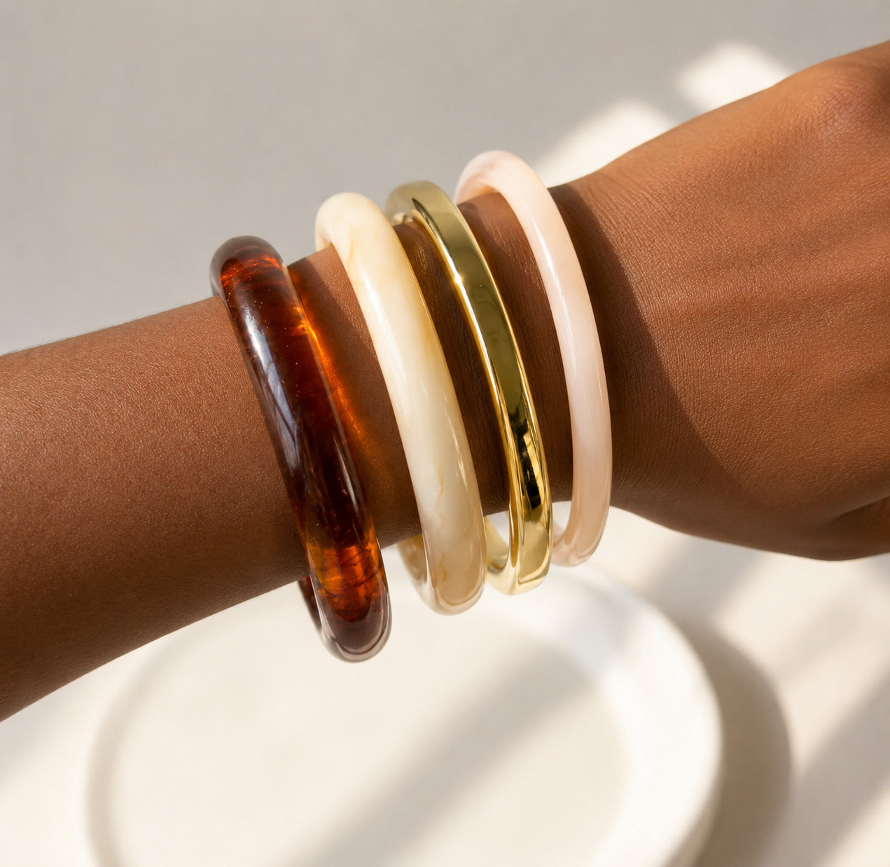 Four bangle bracelets stacked on a person's wrist, including a translucent amber-brown bangle, a cream bangle, a polished gold bangle, and a pale pink bangle.
