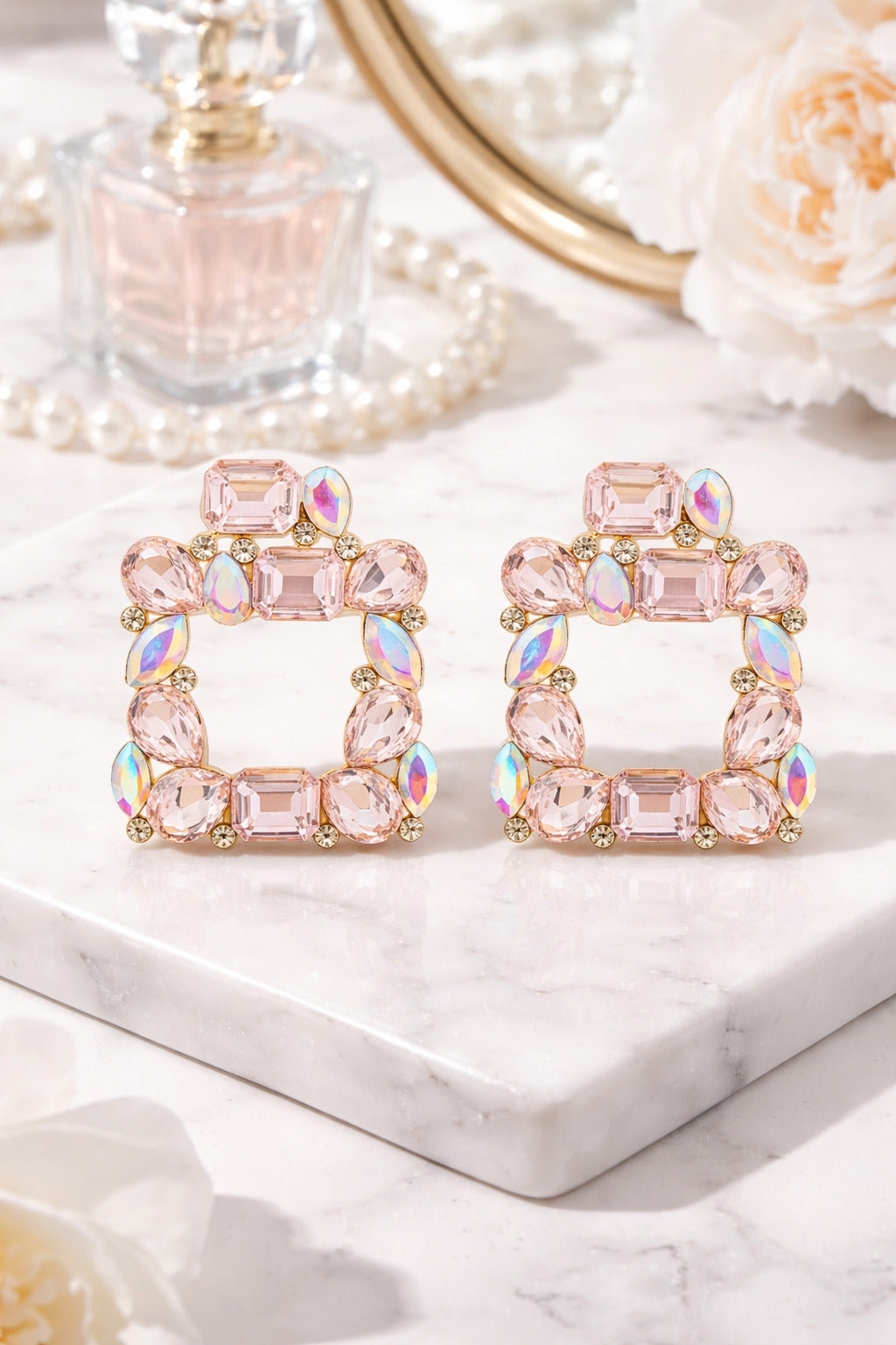 Pair of pink gemstone earrings on a marble surface with a mirror and perfume bottle in the background.
