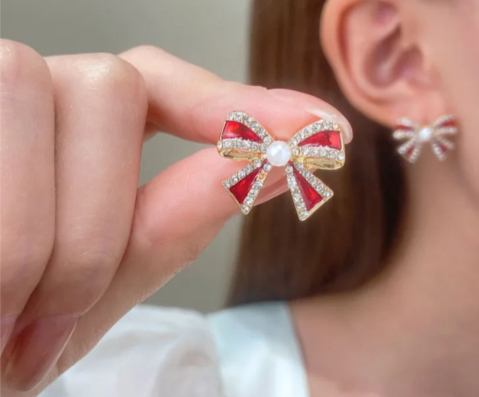 Red bow-shaped earrings with a pearl center held by a hand.
